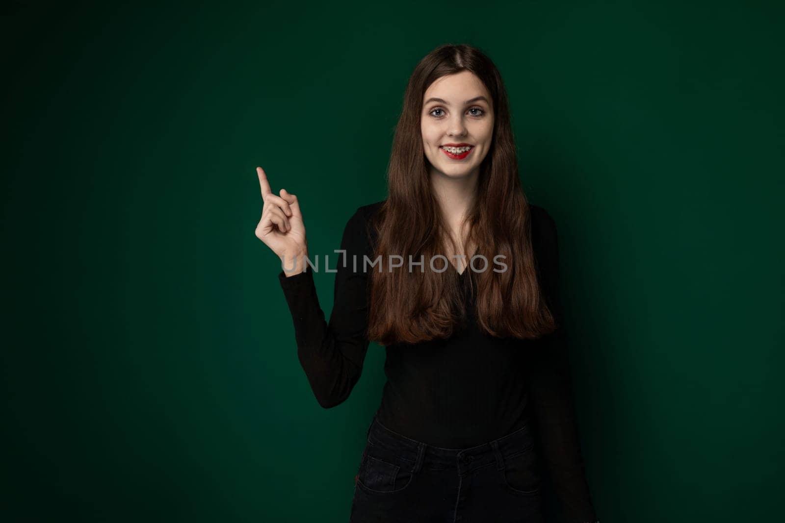 A woman is standing in front of a vibrant green wall, looking directly at the camera with a neutral expression. She is wearing casual attire and appears to be in an urban environment.