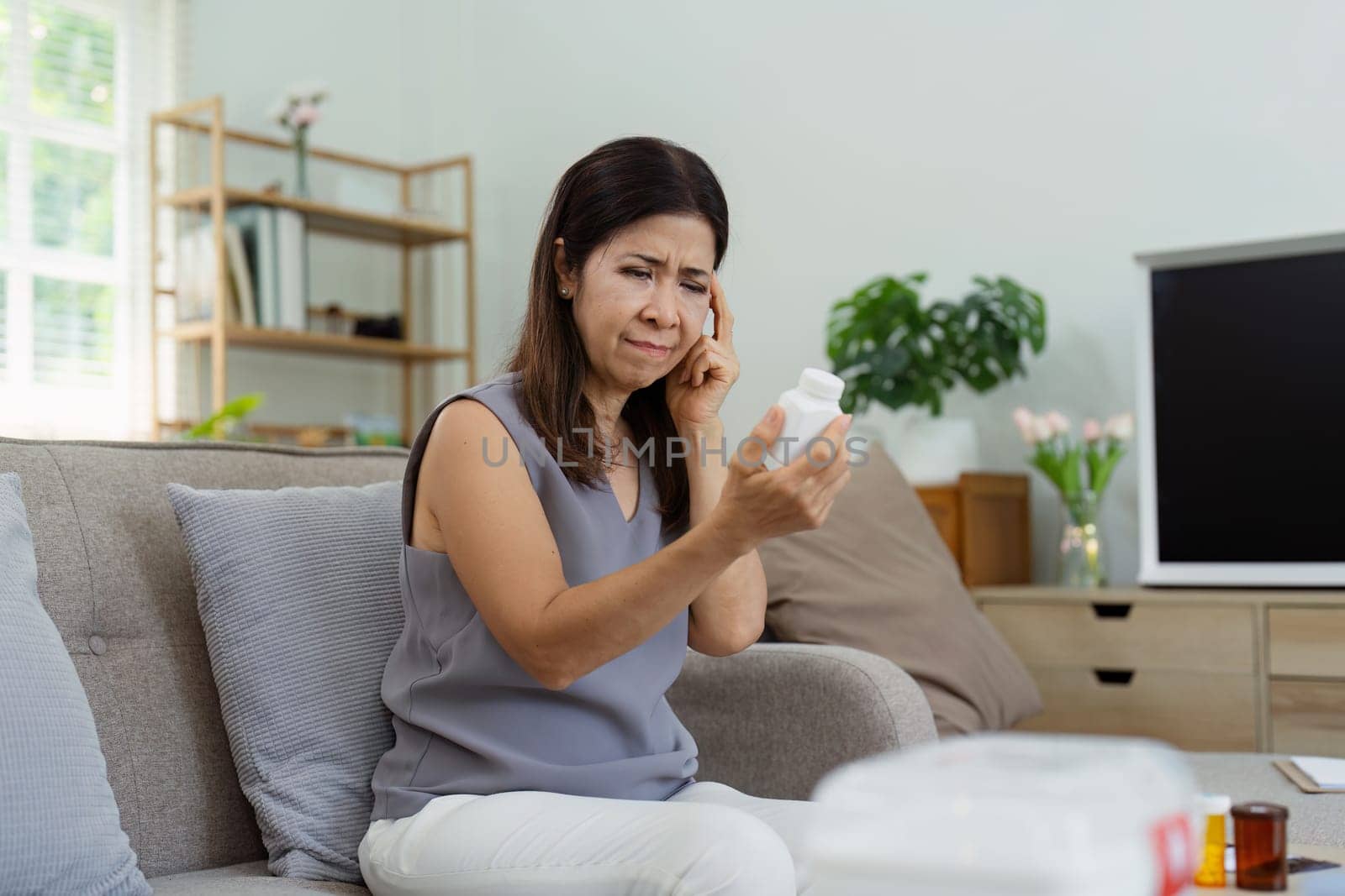 Middle-aged woman carefully reading a medication bottle label while sitting on a comfortable couch in a well-lit, modern living room.