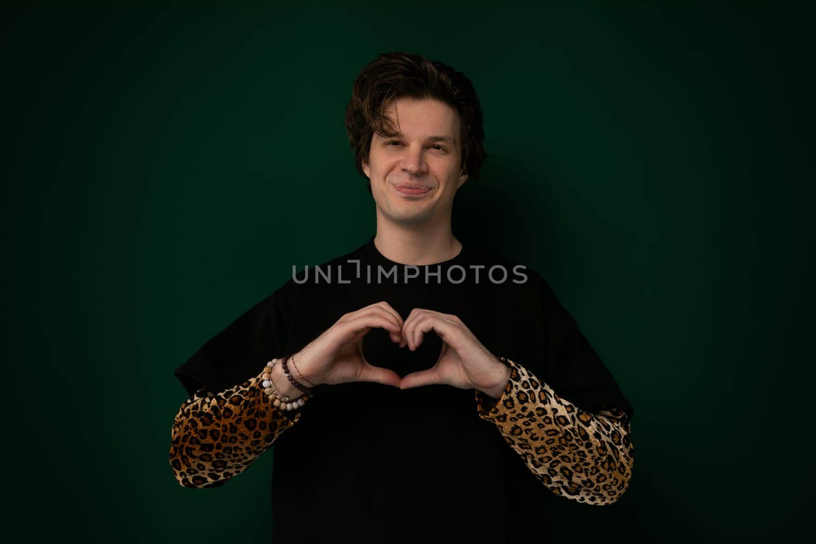 A man is standing, forming a heart shape with his hands in front of his chest. The background is blurred, emphasizing the gesture.