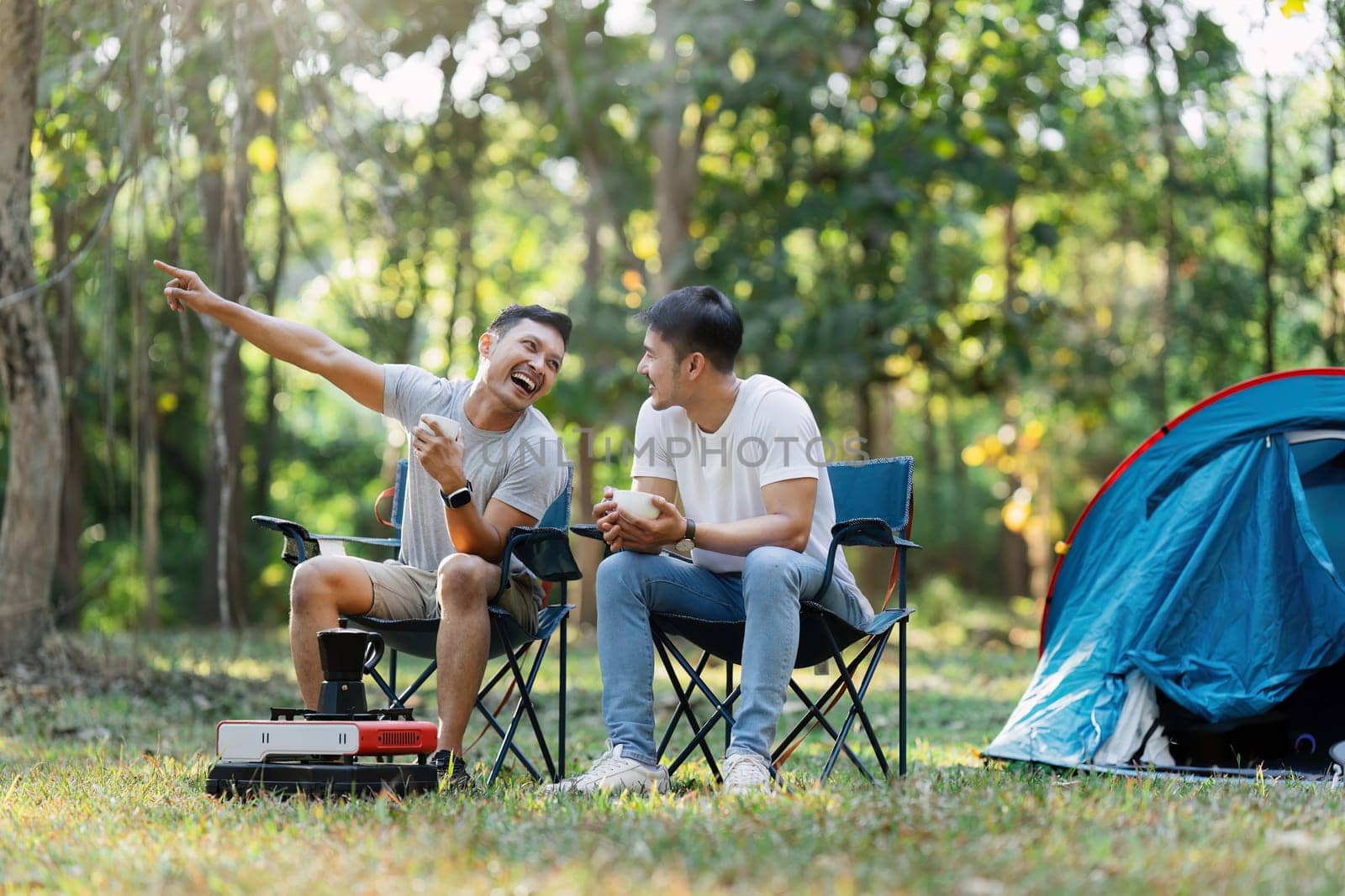 Couple gay lgbt camping in the forest, sitting in chairs, laughing, and drinking coffee with a tent in the background.