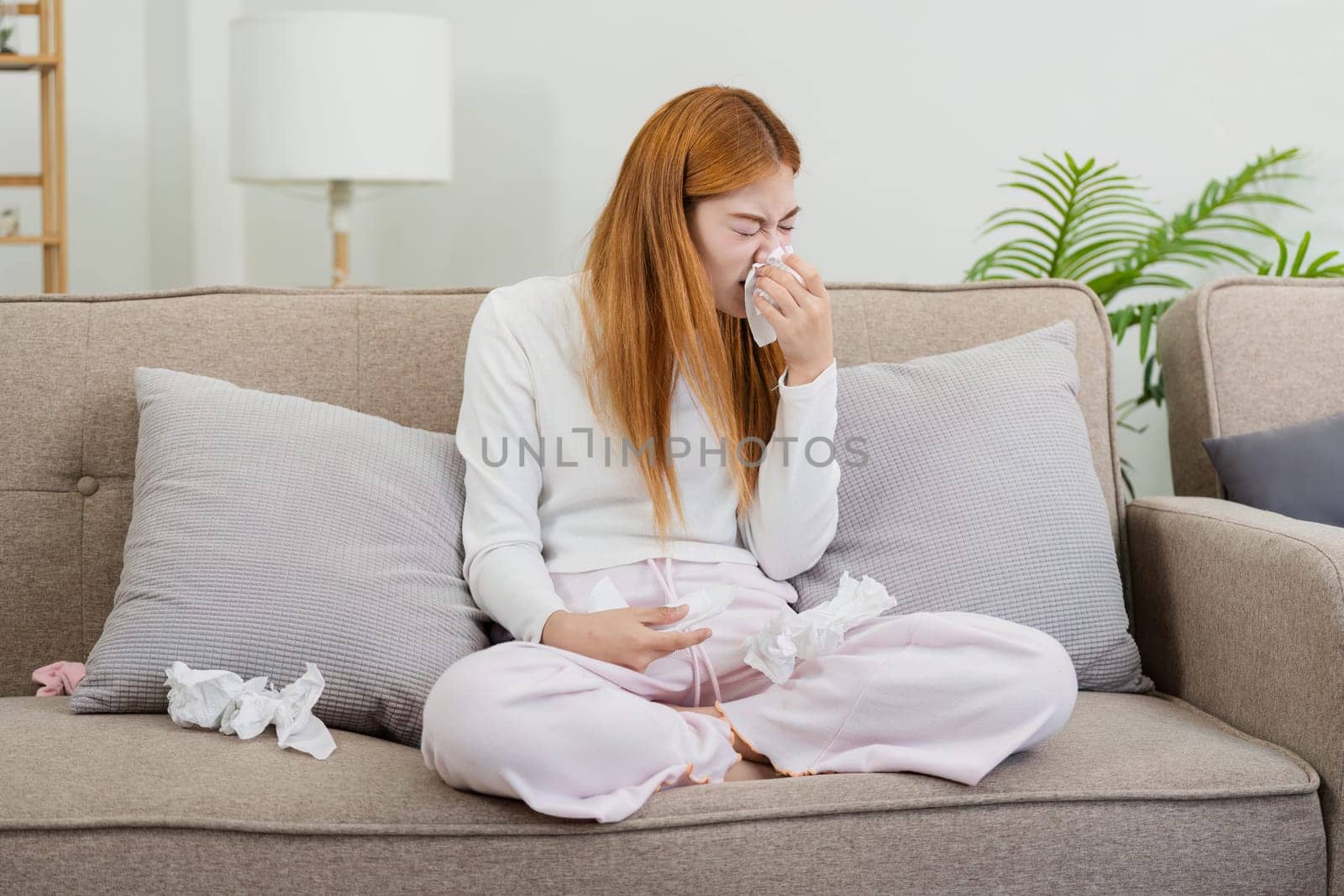 Young woman feeling sick at home, sitting on couch with tissues, blowing nose, and wearing comfortable pajamas in a modern living room.