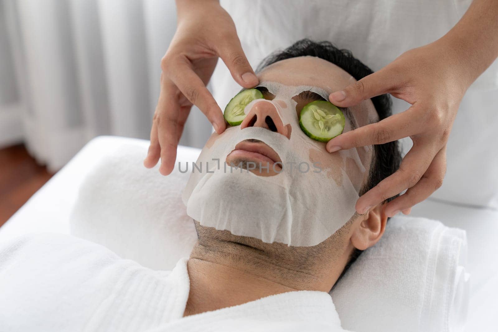 Serene daylight ambiance of spa salon, man customer indulges in rejuvenating with luxurious cucumber facial mask. Facial skincare treatment and beauty care concept. Quiescent