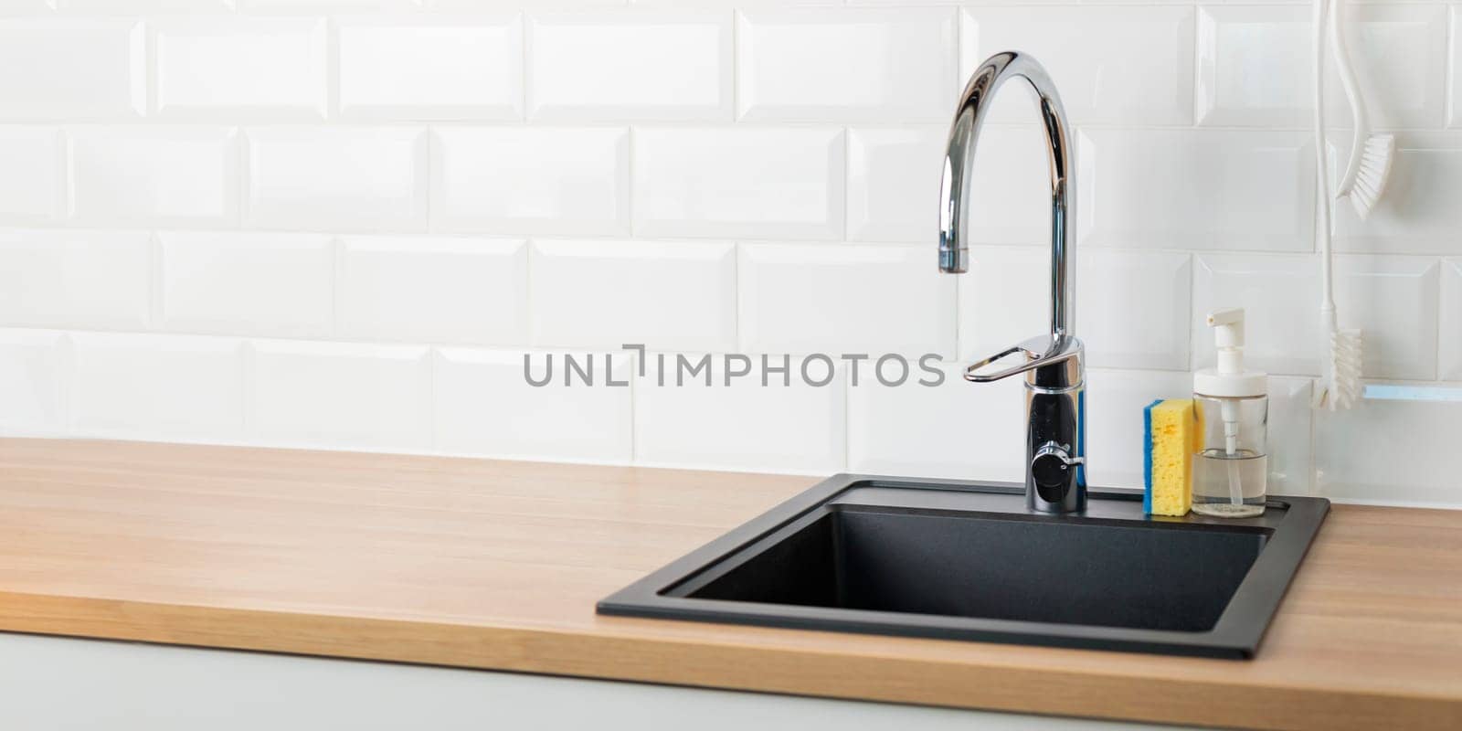 An empty black kitchen sink in a kitchen with a chrome faucet, wooden countertop, and white subway tile backsplash with dishwashing liquid, brush and sponge, banner with copy space