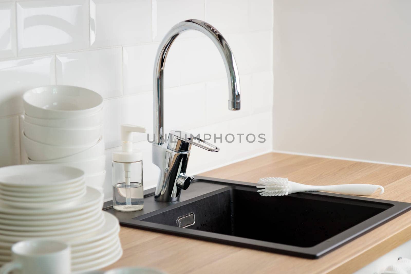 Set of clean washed white dishes near black kitchen sink with a chrome faucet on wooden counter, and white subway tile backsplash with dishwashing liquid and brush..