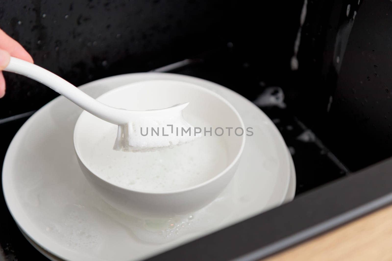 Washing of white plates dishes with brush in the black sink, tableware cleaning with soap foam concept