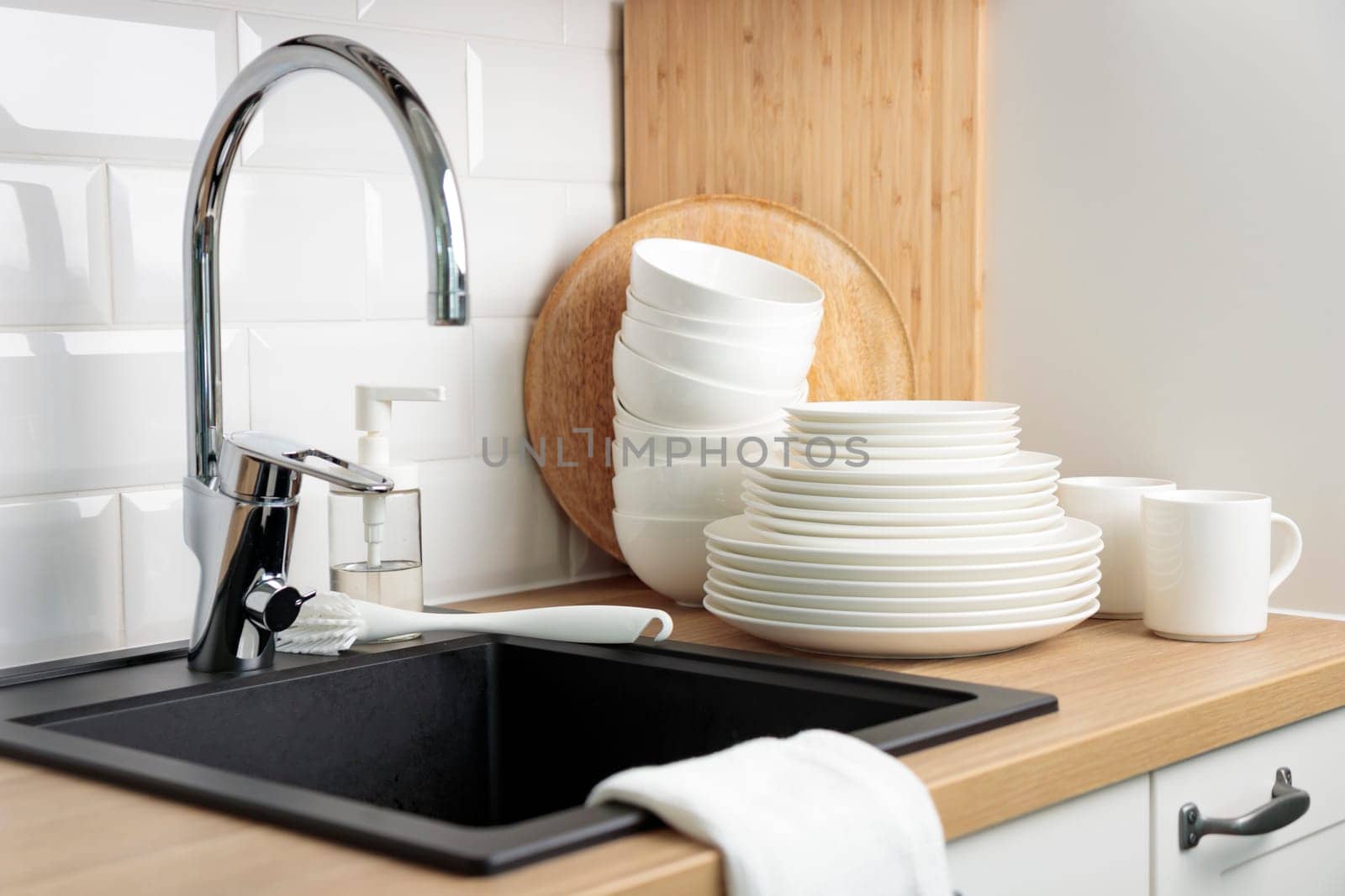 Clean washed tableware. Concept of washing dishes. Dishwashing liquid with brush, plates and cups near black kitchen sink with a chrome faucet on wooden counter and white subway tile backsplash