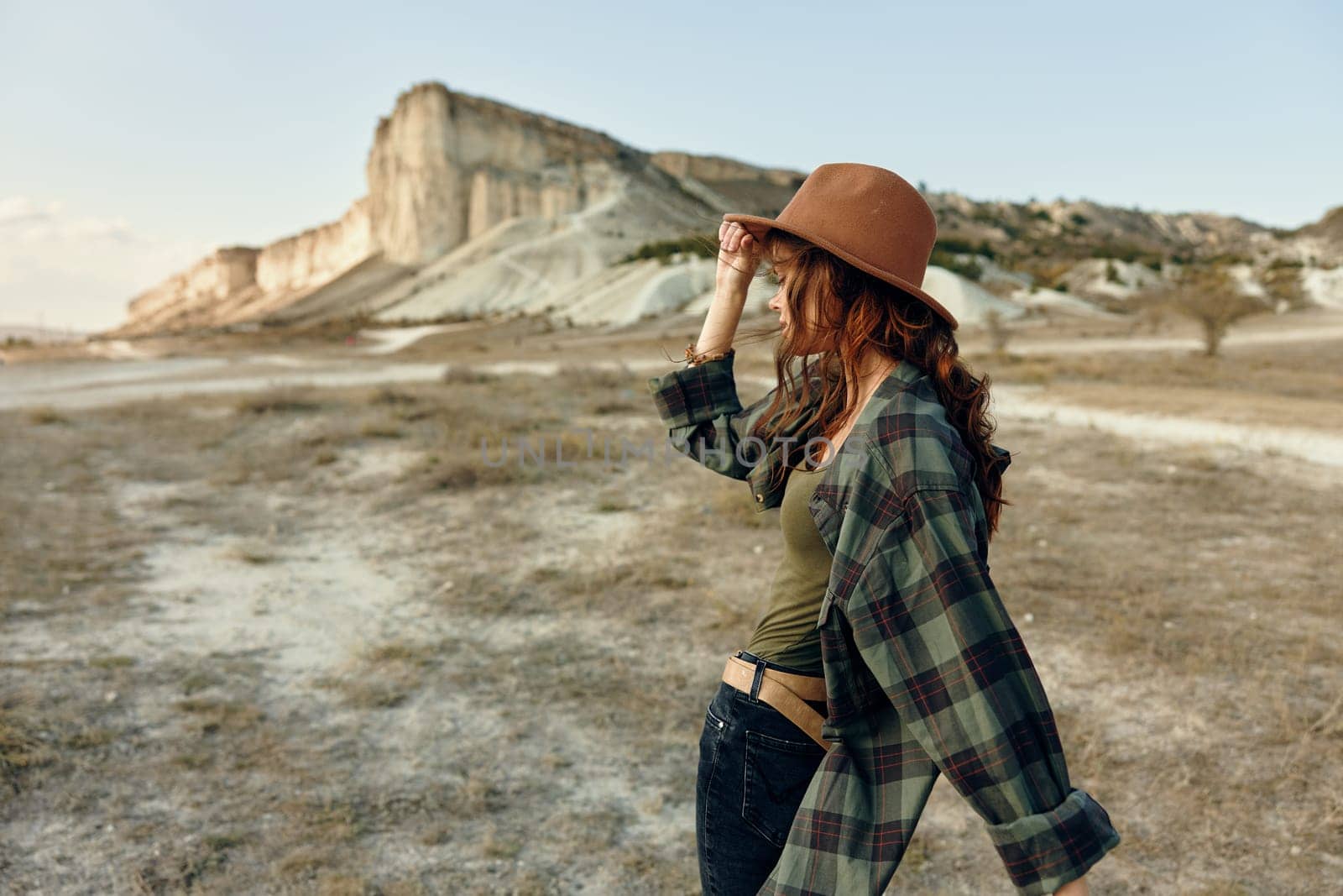 Woman in plaid shirt and hat hiking through desert landscape with mountains in background