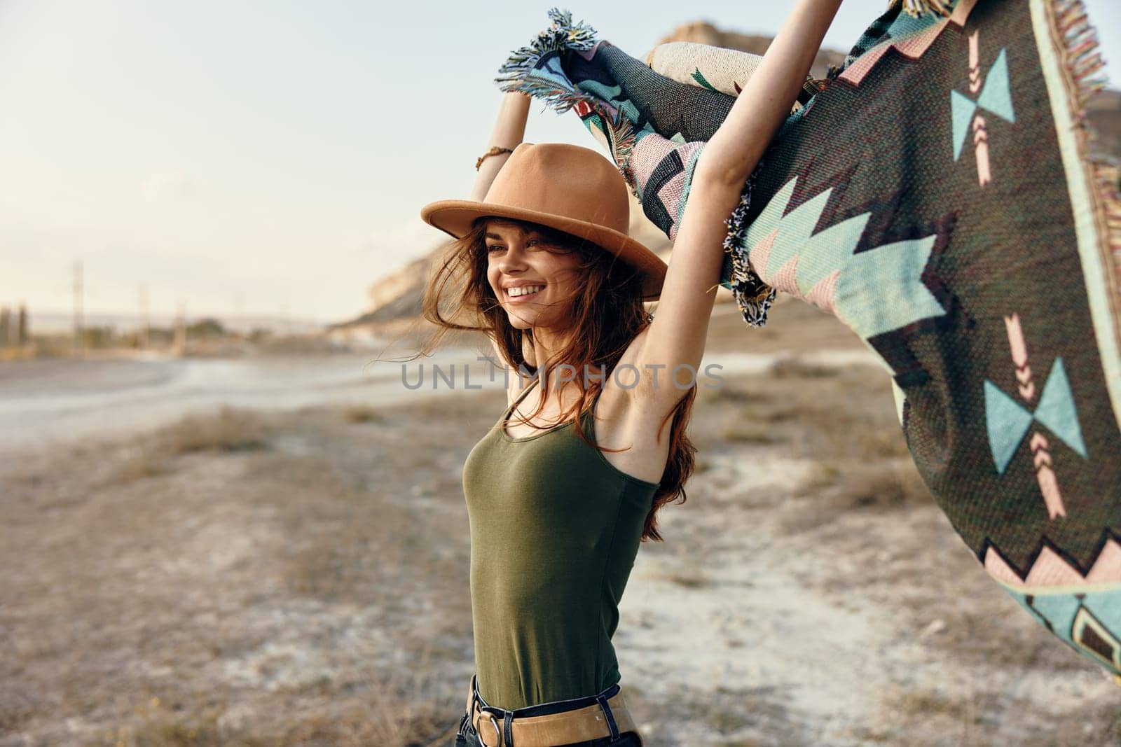 Woman in hat and tank top rejoicing in desert with arms raised holding blanket by Vichizh