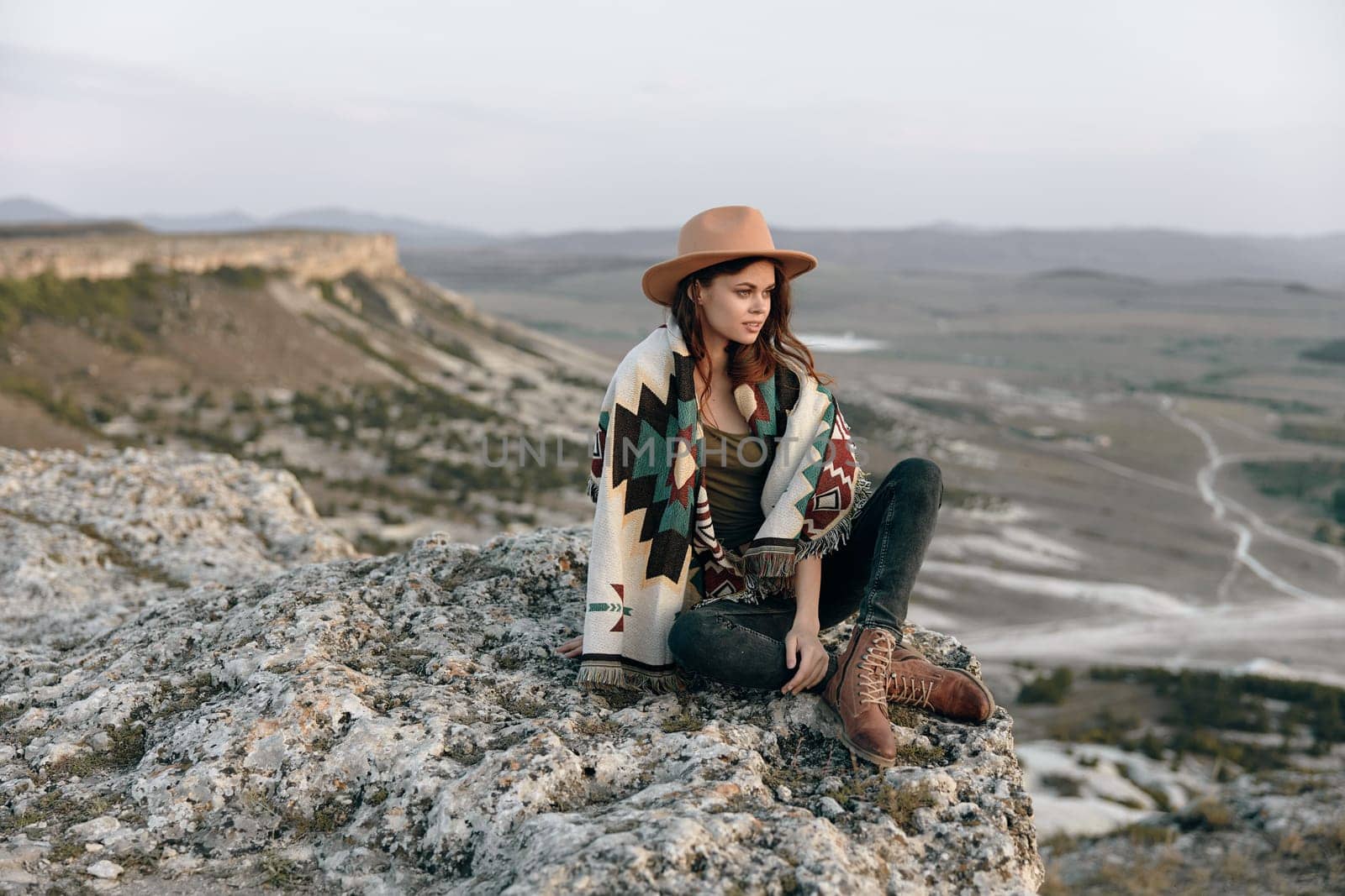 serene woman in the mountains sitting on a rock with a hat and blanket on her shoulders