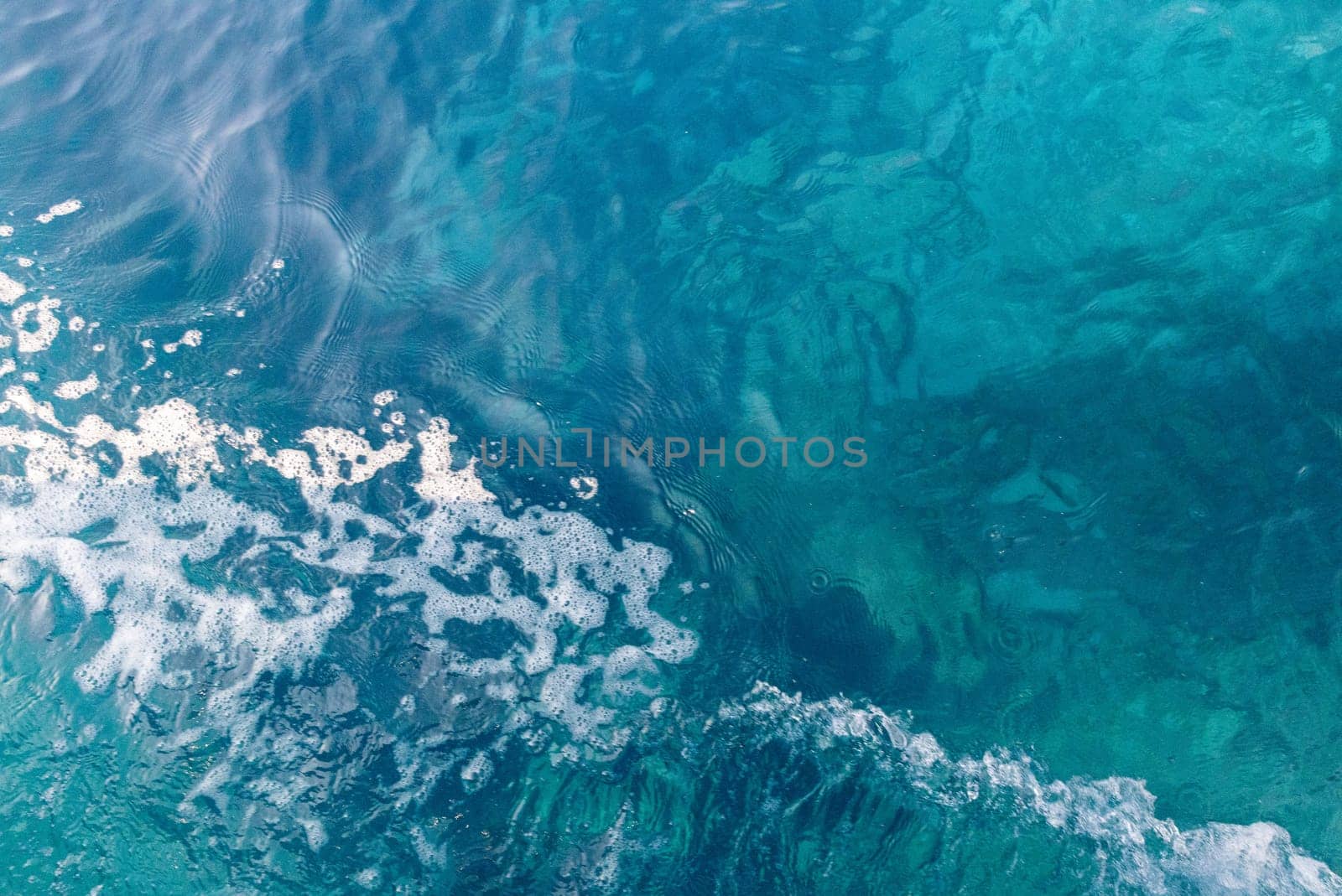 Beautiful view of blue colored water with white foamy wave on the left on a sunny summer day, close up view from above.