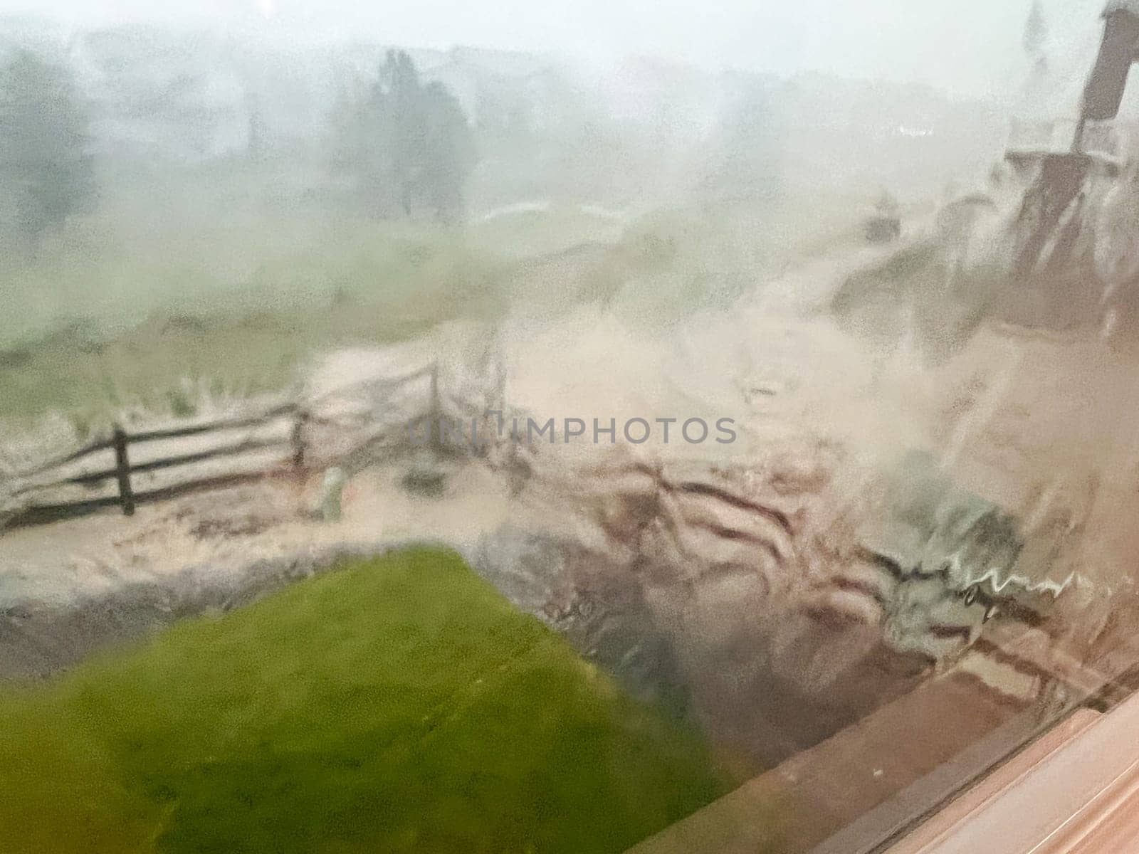 A view through a rain-streaked window capturing a heavy downpour with hail and flash flooding in a suburban backyard. The scene shows blurred fences, trees, and lawn due to the intense rain and hail.