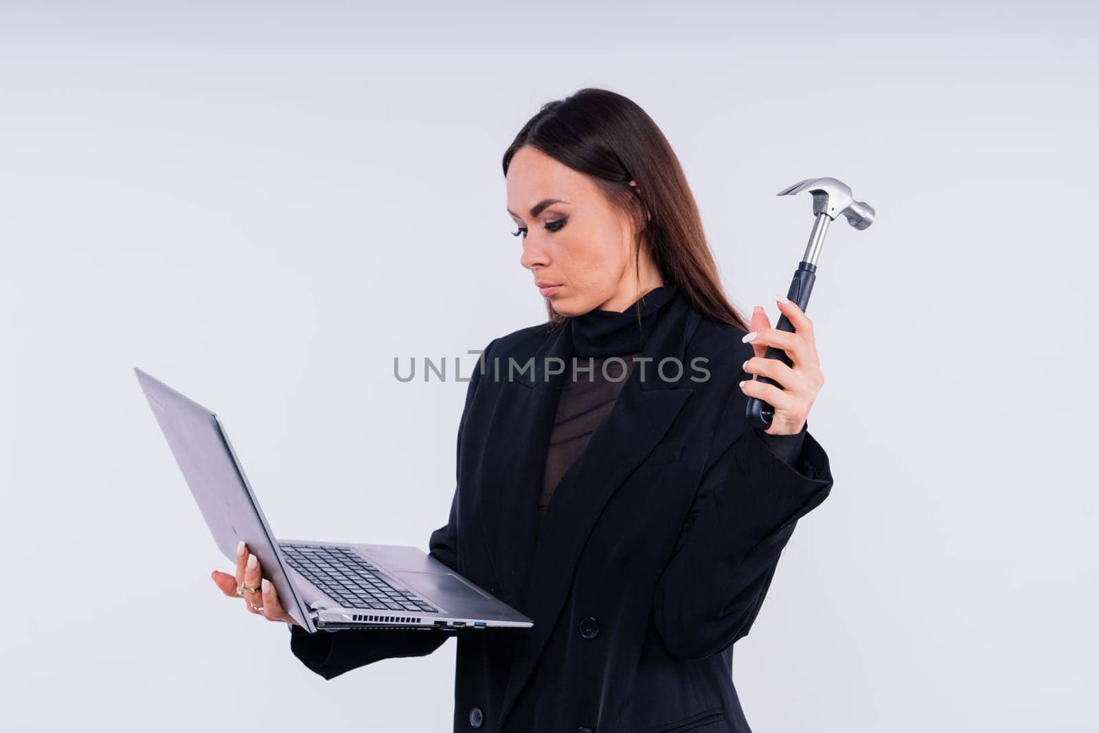 Attractive woman wearing business suit with computer angry facial expression holding hammer