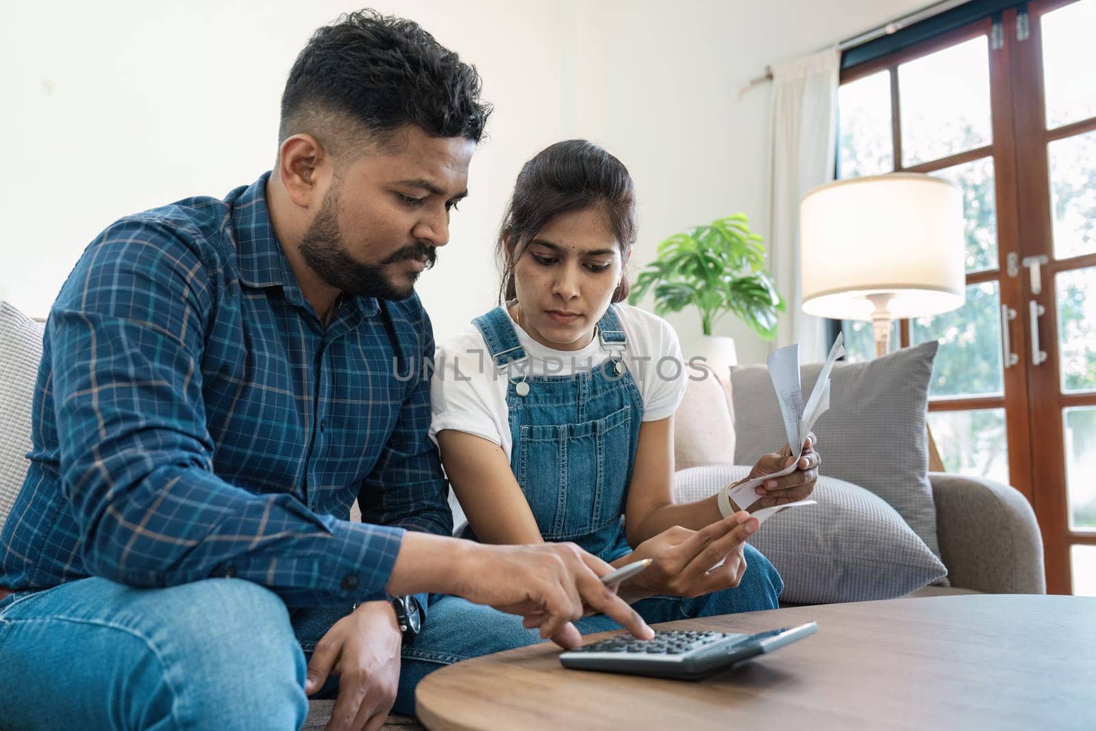 Indian couple sitting in their living room, using a calculator and holding bills, planning their finances together.