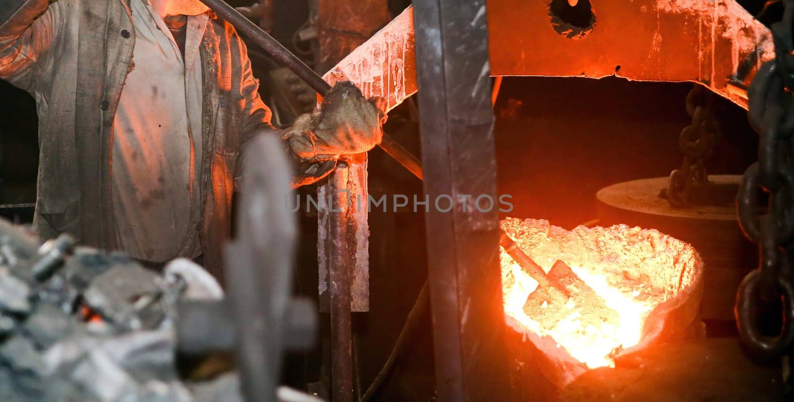 Steel mill worker near furnace with sparks, wearing protective gear, using metal rod to check molten metal, background filled with molten metal and sparks, hard hat and safety glasses worn.