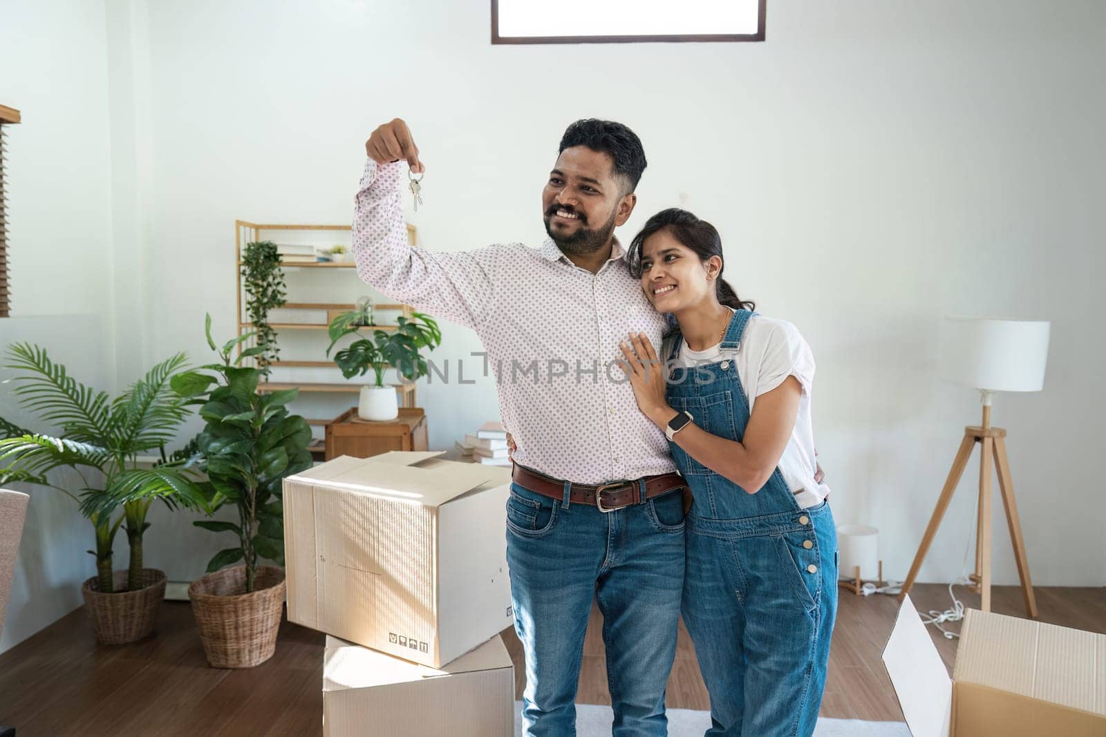 Indian couple moving into a new home, holding keys, surrounded by cardboard boxes and houseplants, celebrating new beginnings in a modern living room.