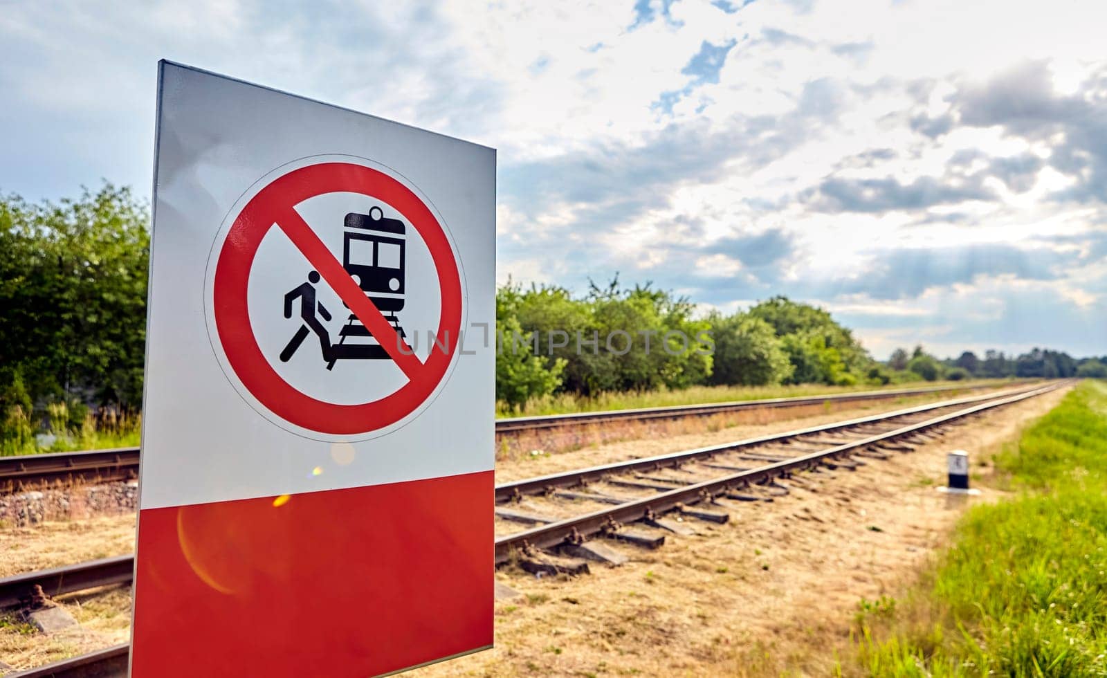 A concrete railway track under a sunny sky with a vivid prohibition sign displaying a train and a person, emphasizing no crossing. The scene captures rail safety on a lovely day.