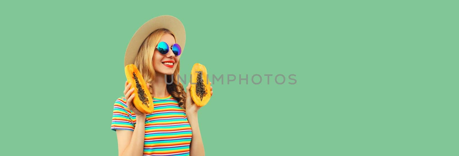 Summer portrait of happy cheerful young woman having fun with fresh papaya fruits wearing straw hat, sunglasses on green background, copy space