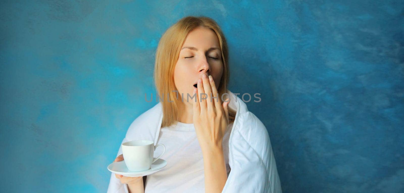 Sleepy lazy young woman yawns waking up after sleeping wrapped in white soft comfortable blanket holds cup of coffee in morning on blue background at home