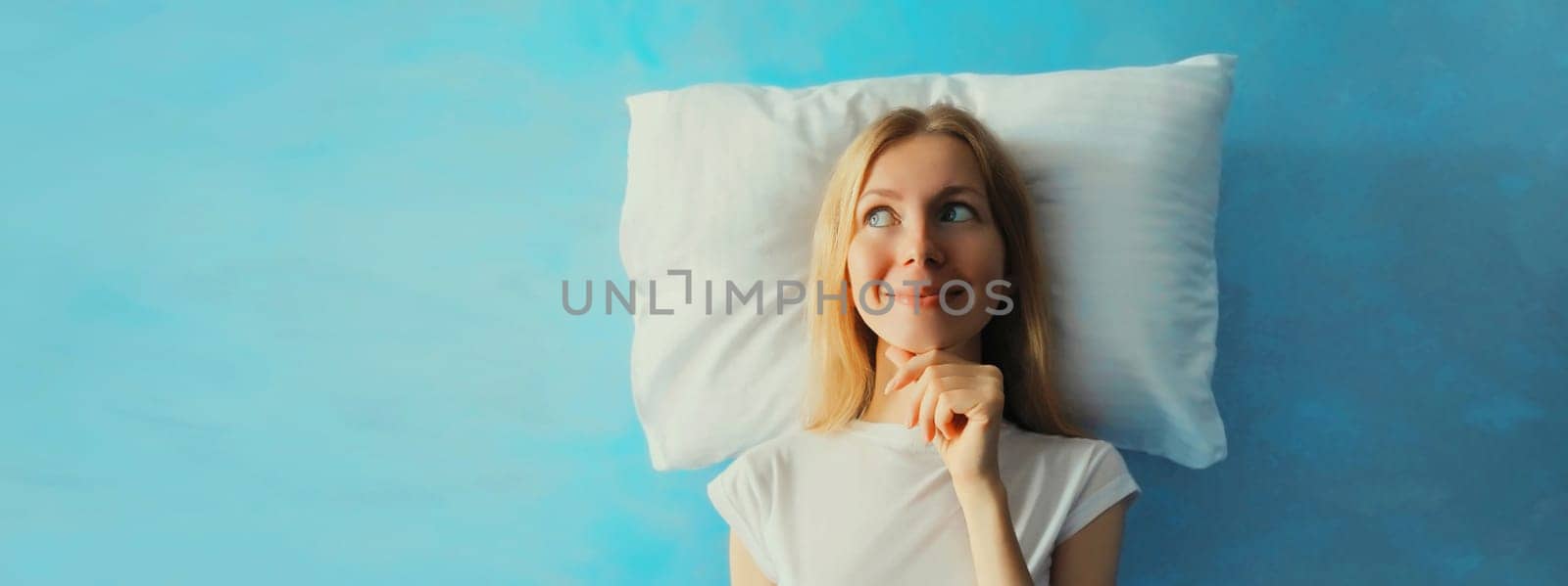 Healthy sleep, happy relaxed sleepy young woman on white soft comfortable pillow thinking looks away imagining she lying on the bed on blue sky with clouds background