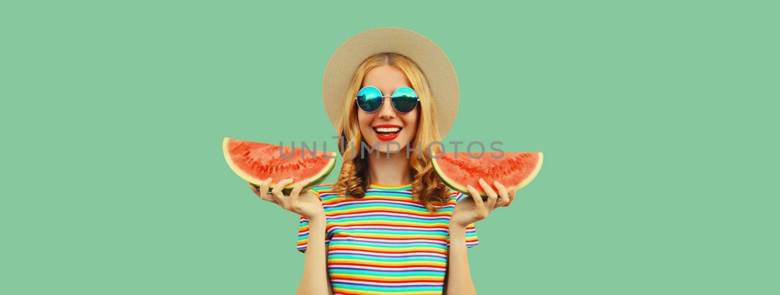 Summer portrait of happy young woman eating fresh slice of watermelon in straw tourist hat, sunglasses on green studio background