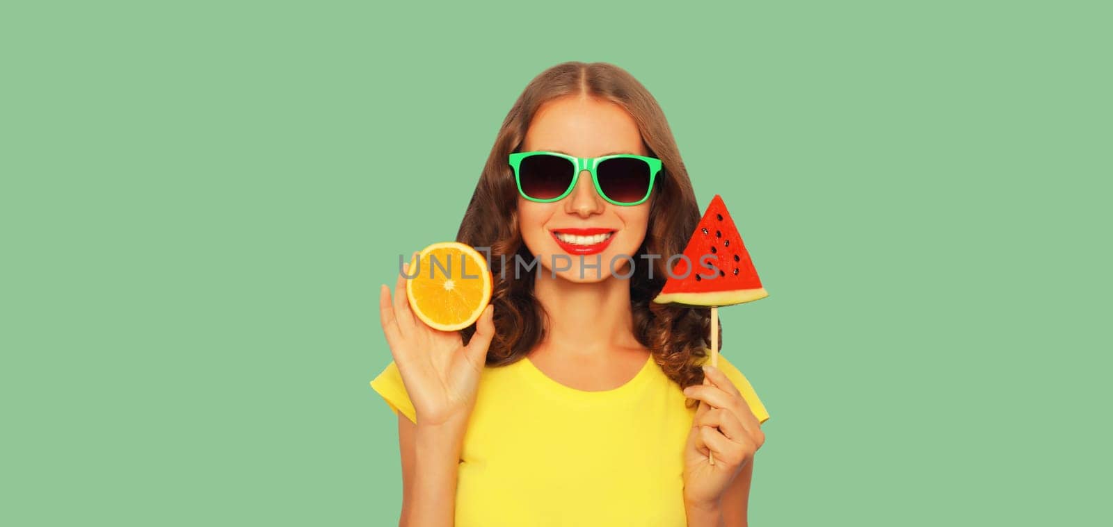 Summer portrait of happy smiling young woman with orange fruit and lollipop or ice cream shaped slice of watermelon wearing glasses on green background