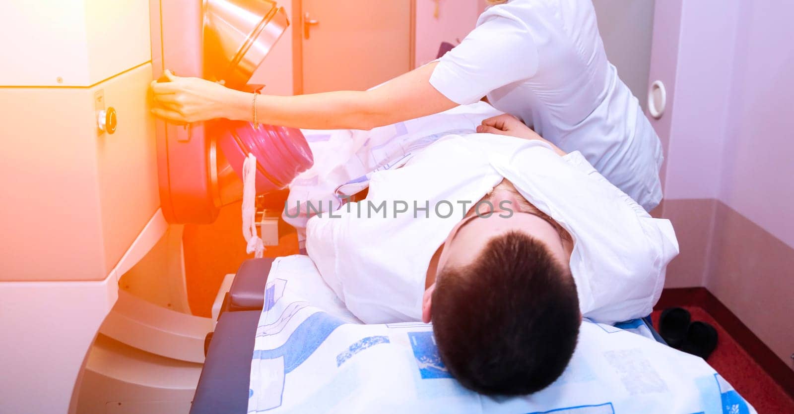 A young male patient is lying on a table getting a medical procedure done. A nurse is assisting him. The patient is wearing a white shirt and the nurse is wearing a white lab coat.