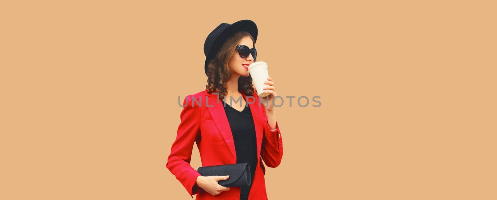 Stylish elegant woman posing in business suit, red blazer jacket, black round hat with handbag clutch looking away isolated on brown studio background