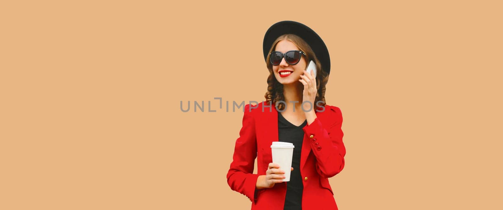 Beautiful lady woman calling on smartphone in red business suit, black hat isolated on brown studio background, copy space for advertising text