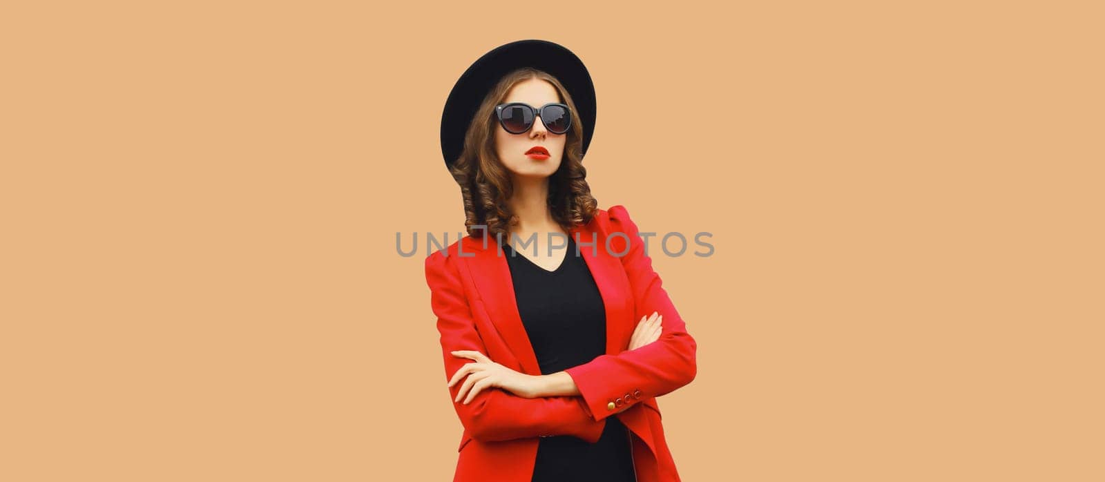 Stylish elegant woman posing in business suit, red blazer jacket and black round hat on brown studio background