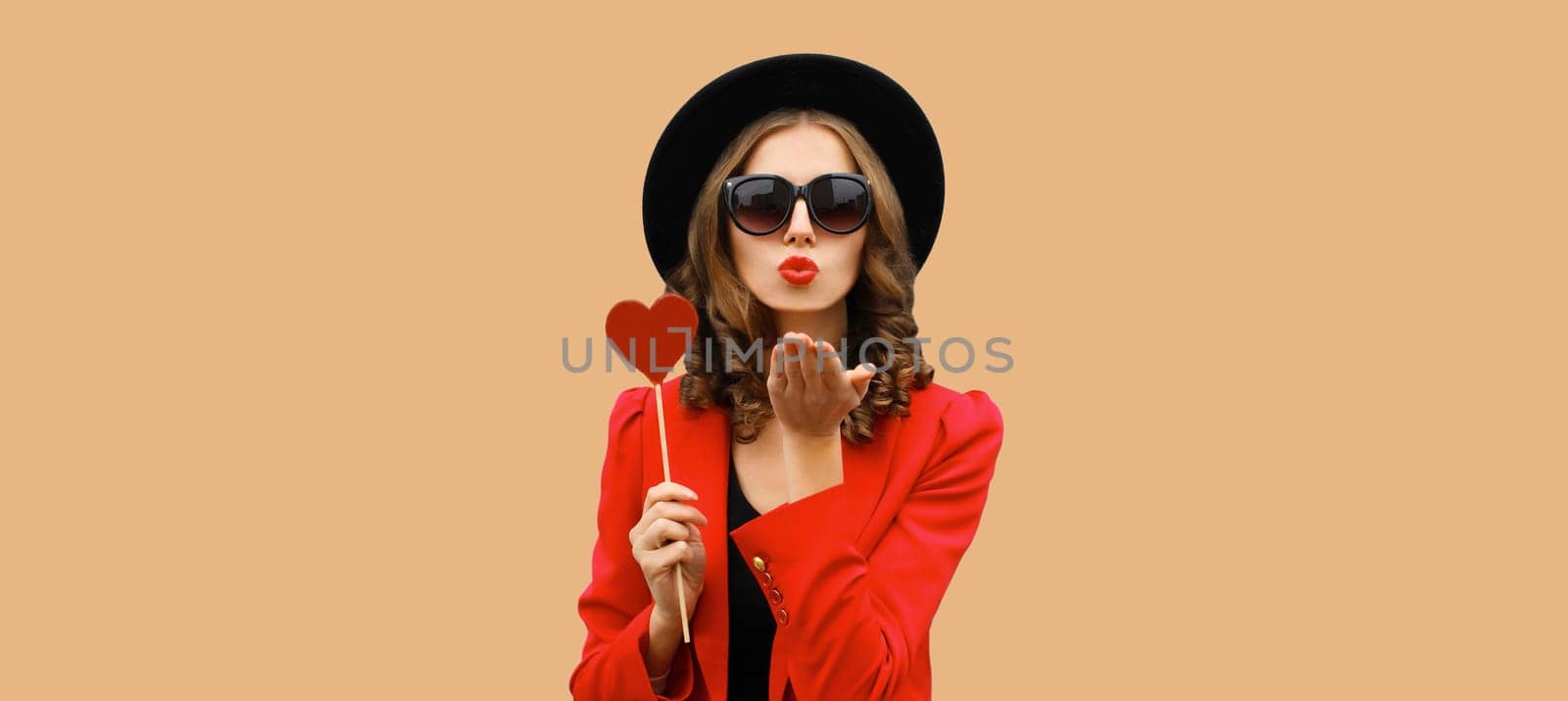 Beautiful stylish woman blowing kiss with sweet red heart shaped lollipop on stick in black hat by Rohappy