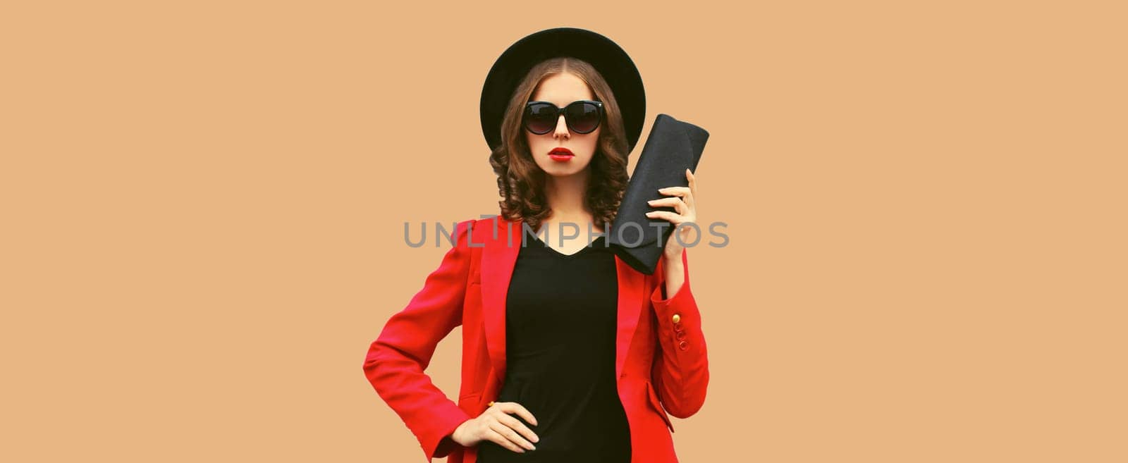 Stylish elegant woman posing in business suit, red blazer jacket, black round hat with handbag clutch posing on brown studio background