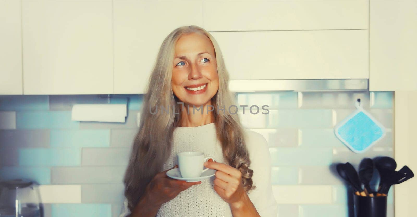 Portrait of happy smiling middle-aged gray-haired woman enjoying hot tasty cup of coffee or tea in the kitchen in early morning at home