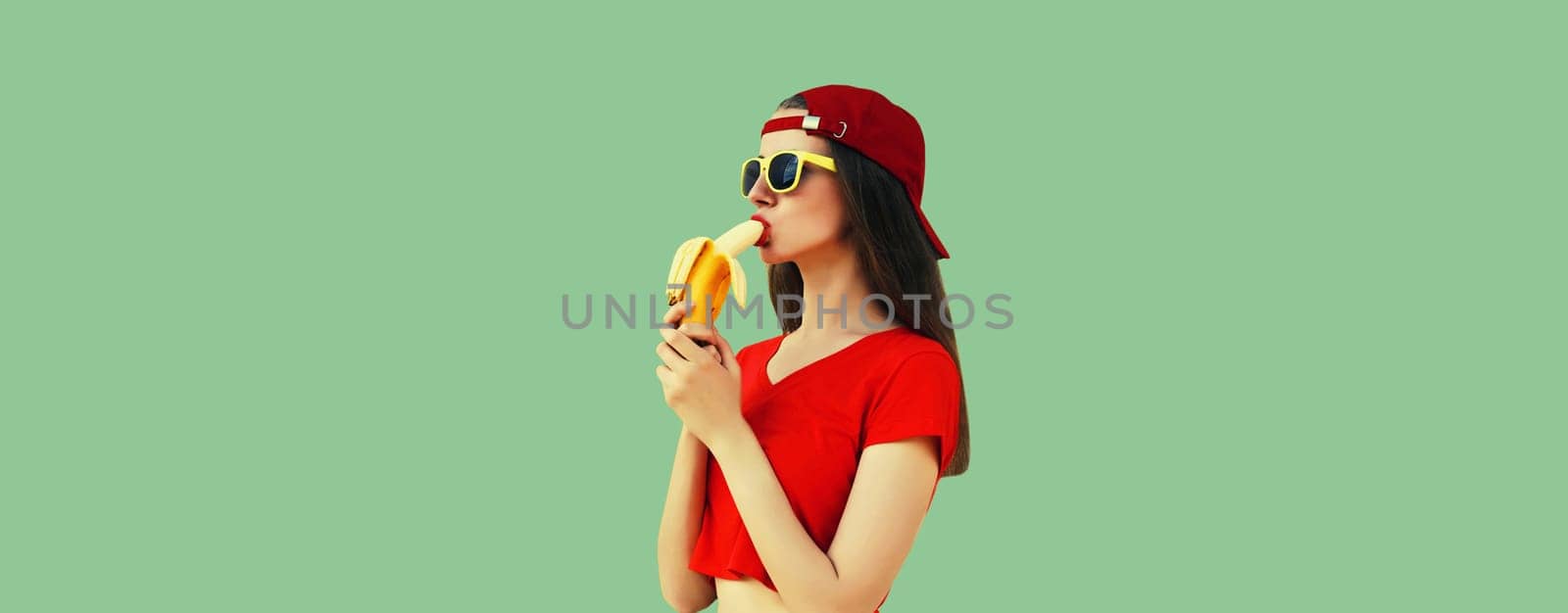 Stylish caucasian young woman eating banana in red baseball cap on green studio background