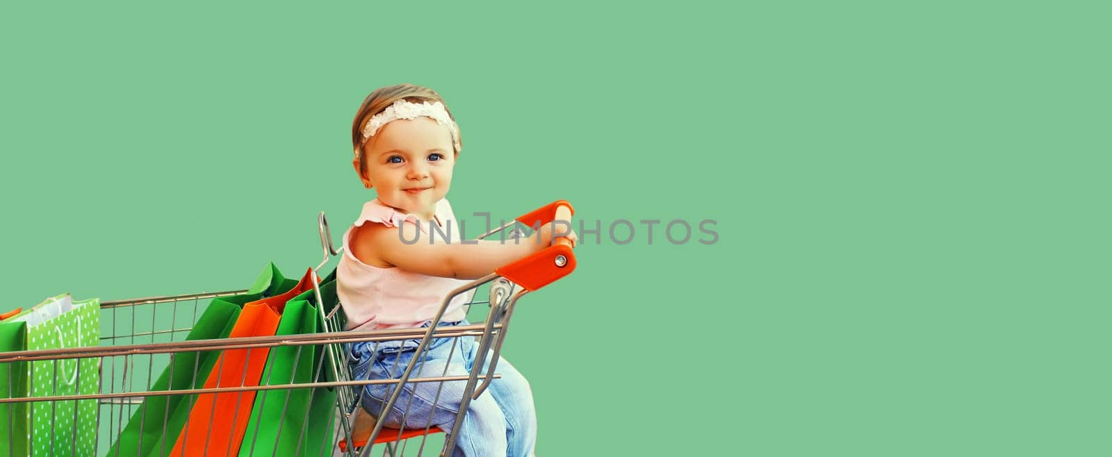 Happy baby with shopping bags sitting in trolley cart on green background by Rohappy