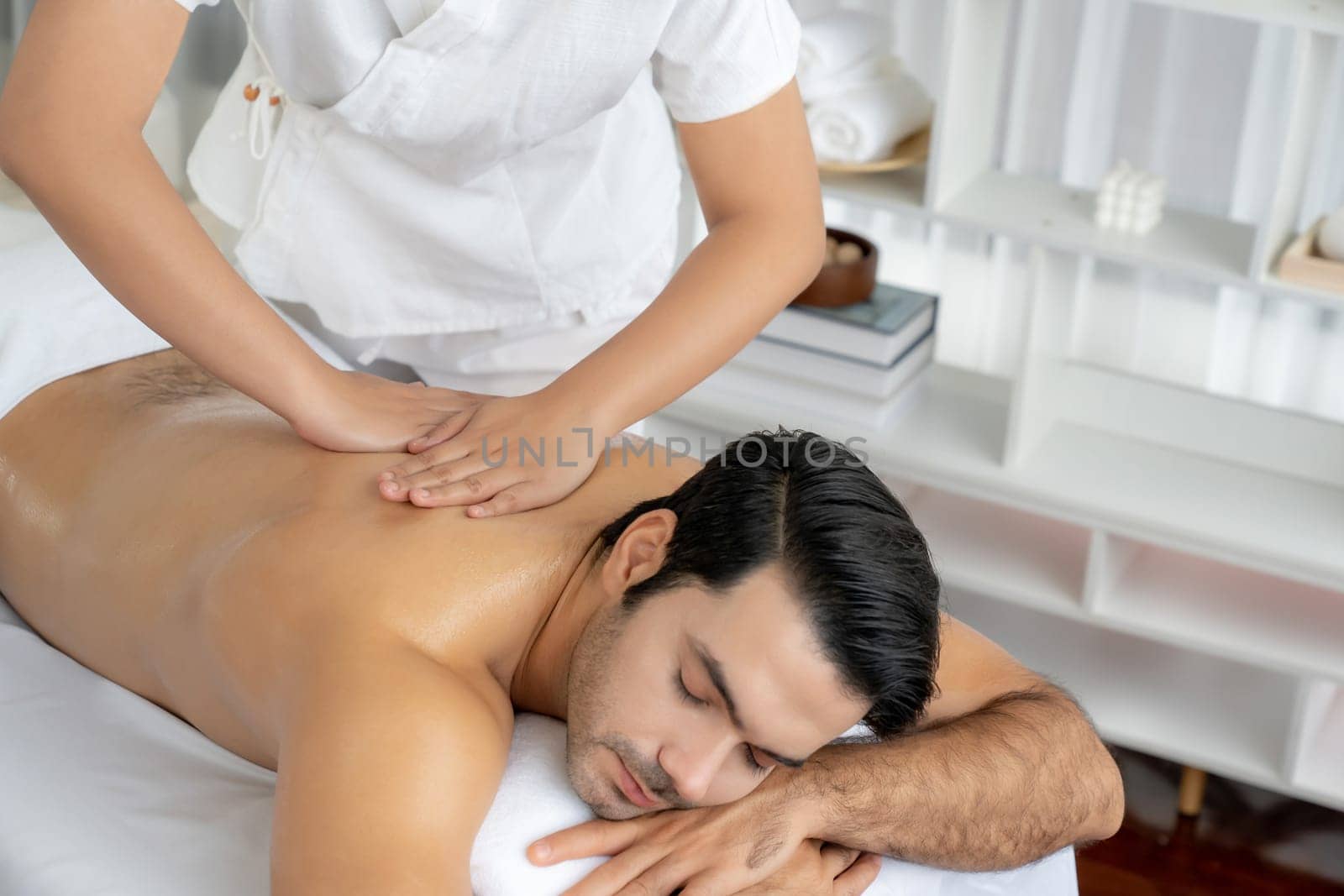 Caucasian man customer enjoying relaxing anti-stress spa massage and pampering with beauty skin recreation leisure in day light ambient salon spa at luxury resort or hotel. Quiescent