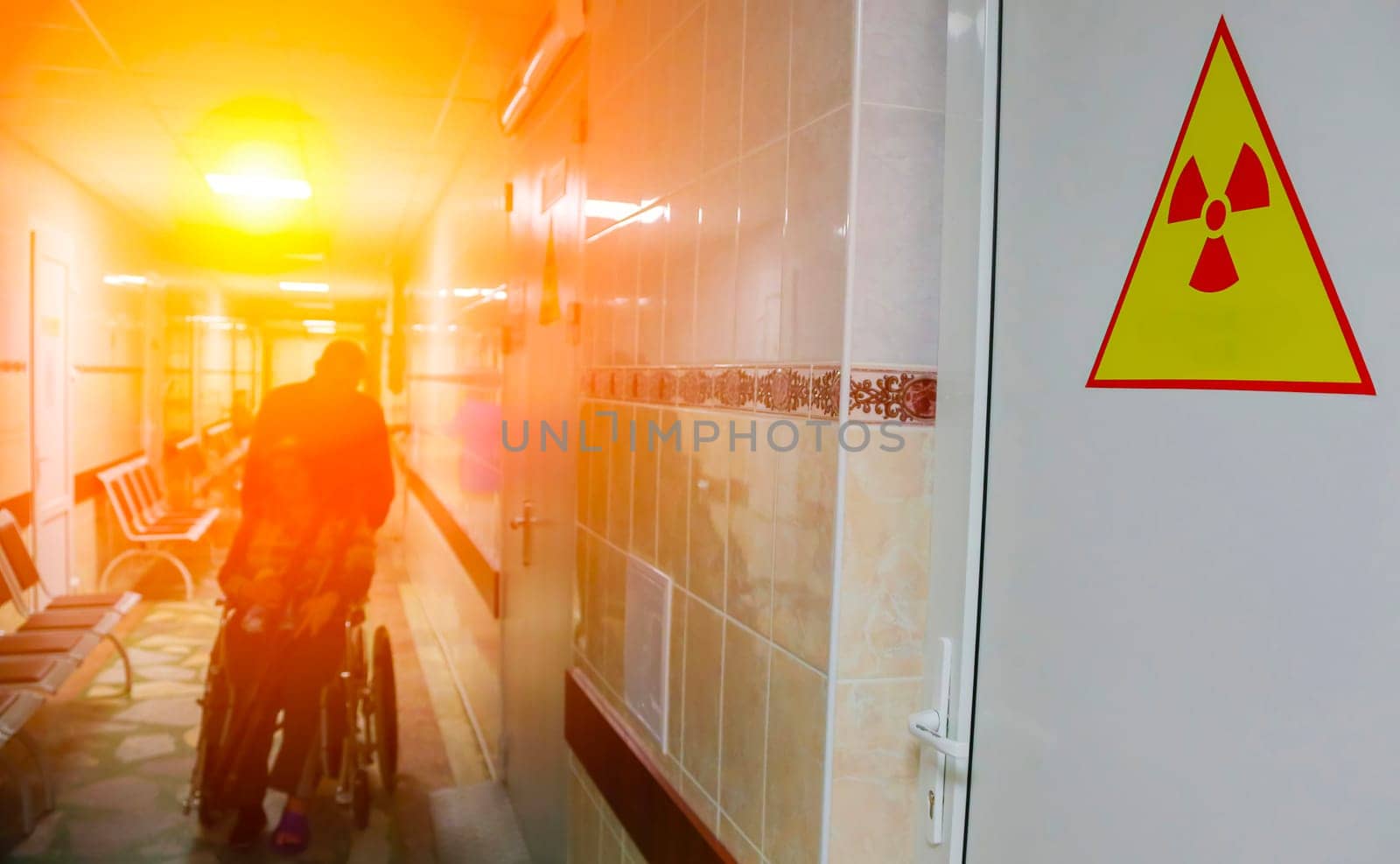 A person in a wheelchair pushes through a hospital corridor with a bright light illuminating the way. A yellow warning sign with a red radiation symbol is prominently displayed on the wall, highlighting the stark contrast between danger and hope.