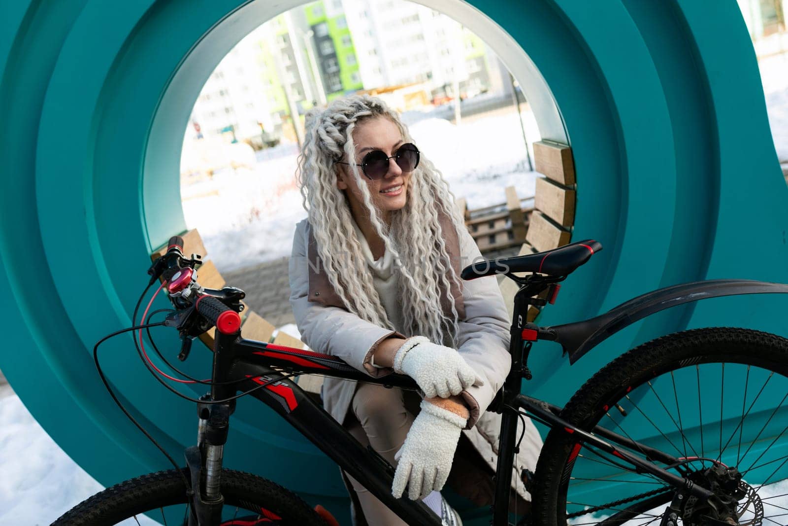 A woman sits on a stationary bike positioned in front of a large circular window. She appears relaxed, looking out through the glass. The surroundings are unseen, focusing on the interaction between the woman and the unique window.