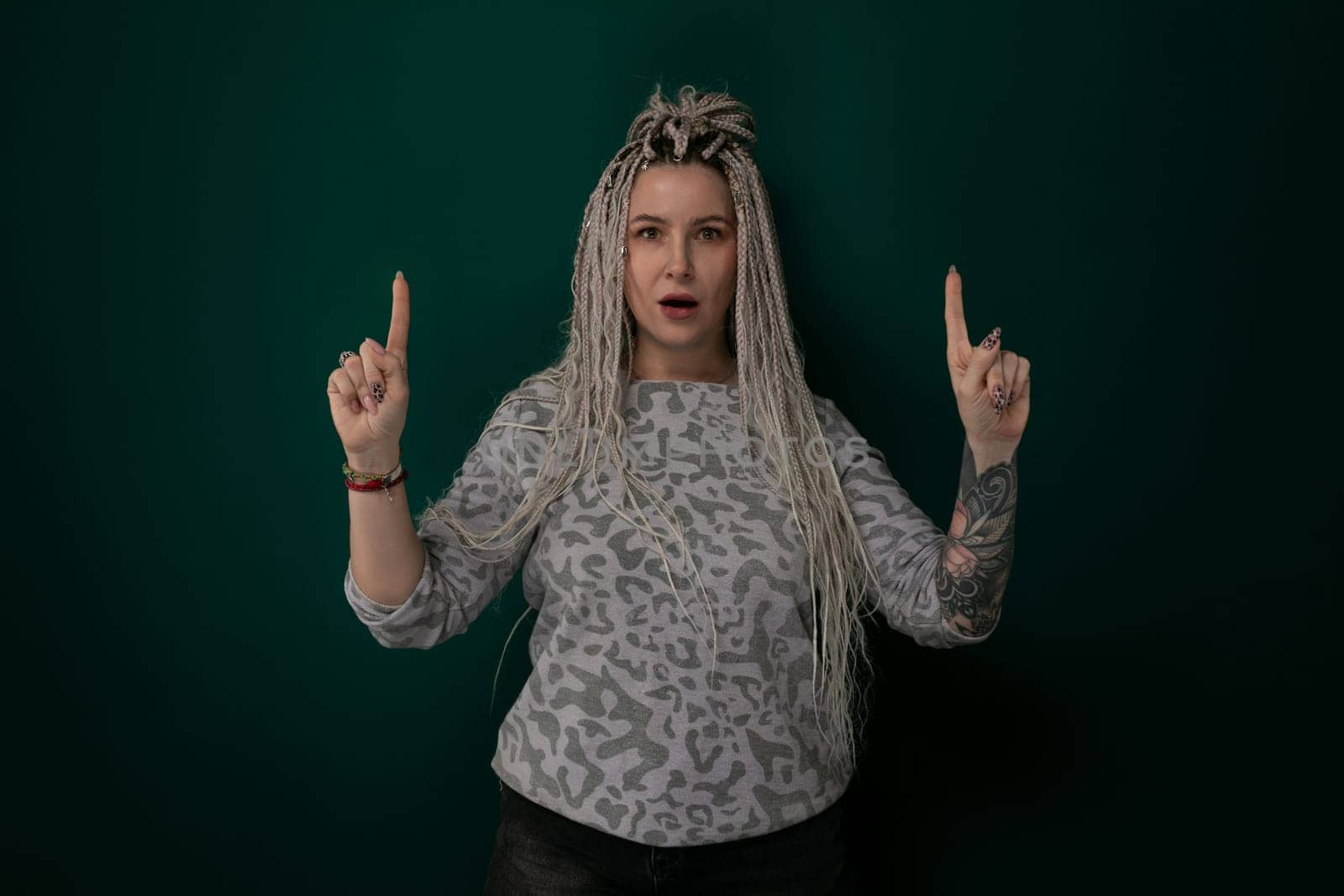 A woman with dreadlocks is standing in front of a vibrant green wall. She appears confident and stylish, with her unique hairstyle drawing attention. The green wall provides a striking backdrop for the scene.