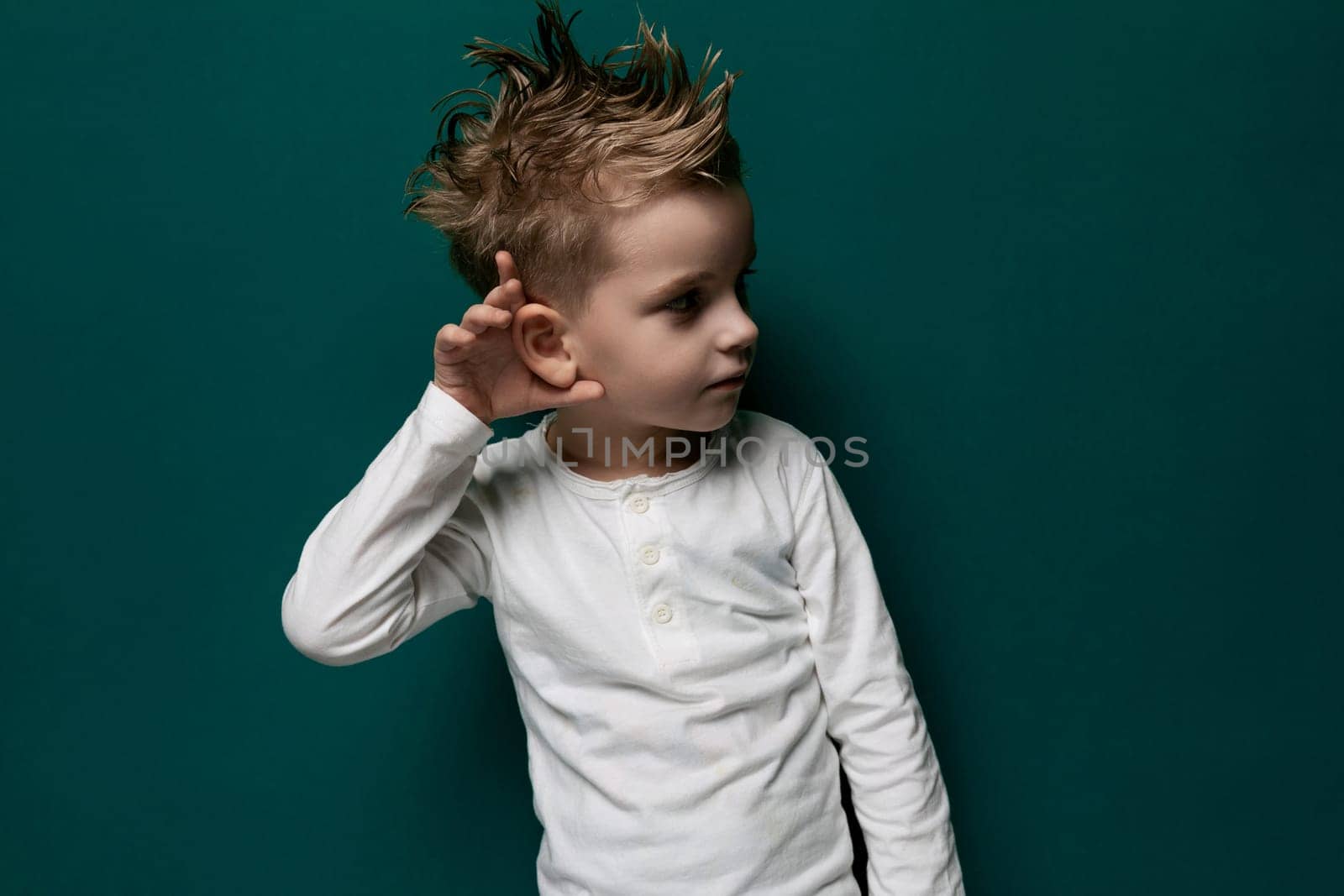 A young boy with a mohawk hairstyle is standing confidently in front of a vibrant green wall. His hands are by his sides, and he is looking directly at the camera.