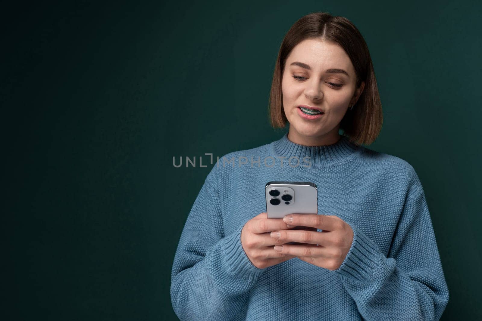 A woman wearing a blue sweater is engrossed in her cell phone screen, her attention focused on the device in her hands as she stands in a room illuminated by natural light.