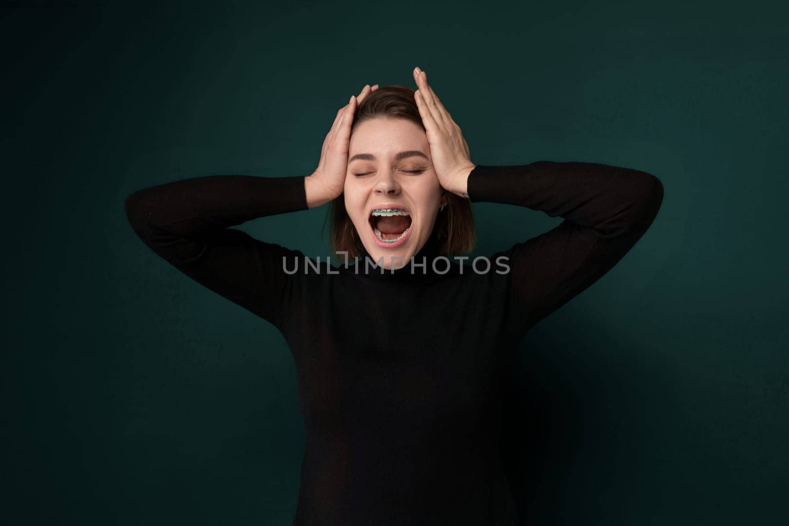 A woman standing with her hands on her head, appearing distressed or in disbelief. She is wearing a white shirt and has long brown hair. The background is a plain wall.