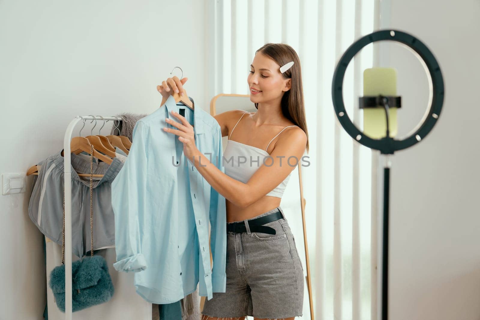 Young woman making fashion video content for social media. Blogger smiles to camera using selfie stick and light ring while making persuasive online clothing sell to audience or followers. Unveiling