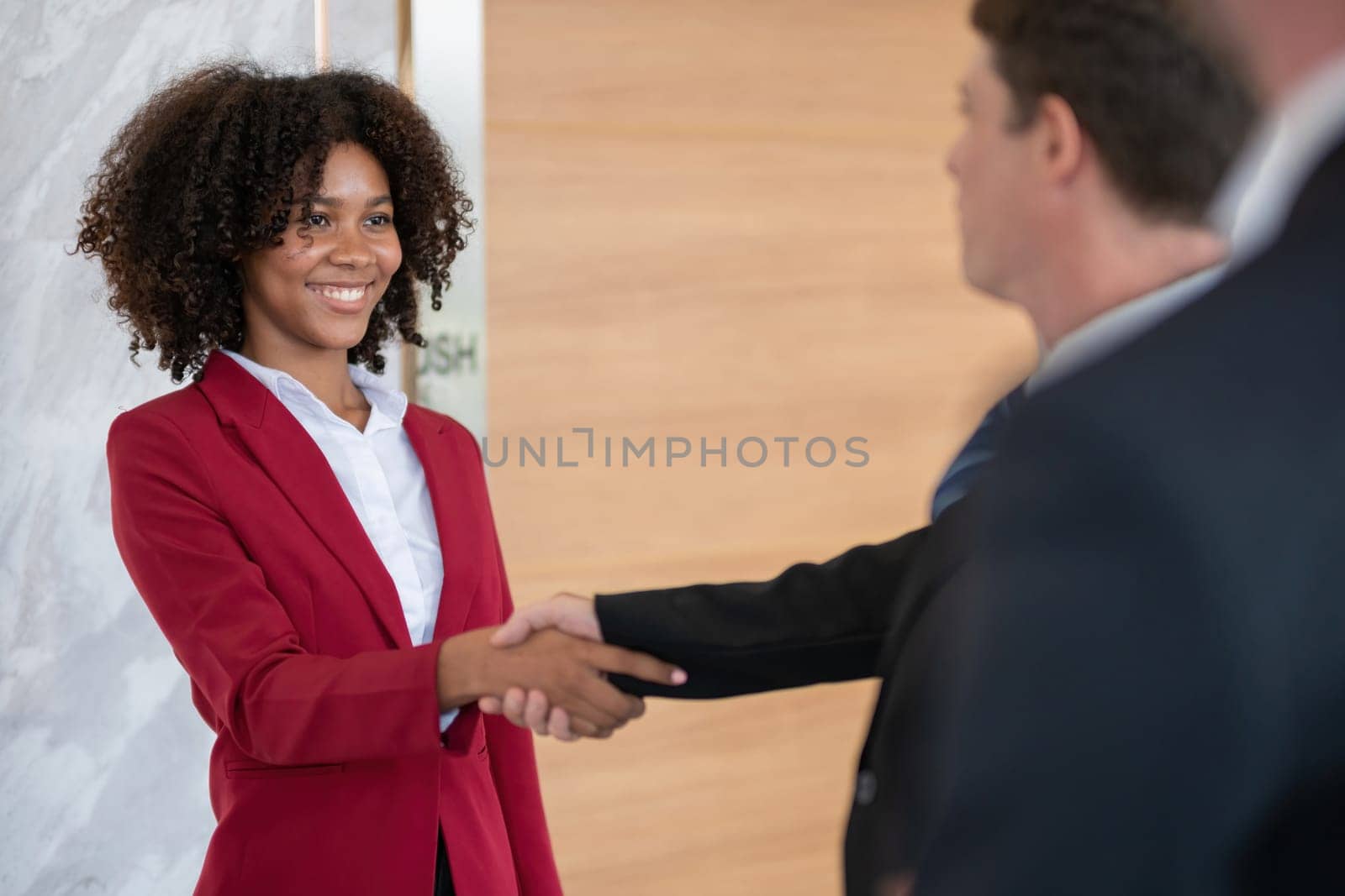 Diverse business team member, a woman, shaking hands with a colleague at an office, smiling and wearing business attire, bright modern office setting