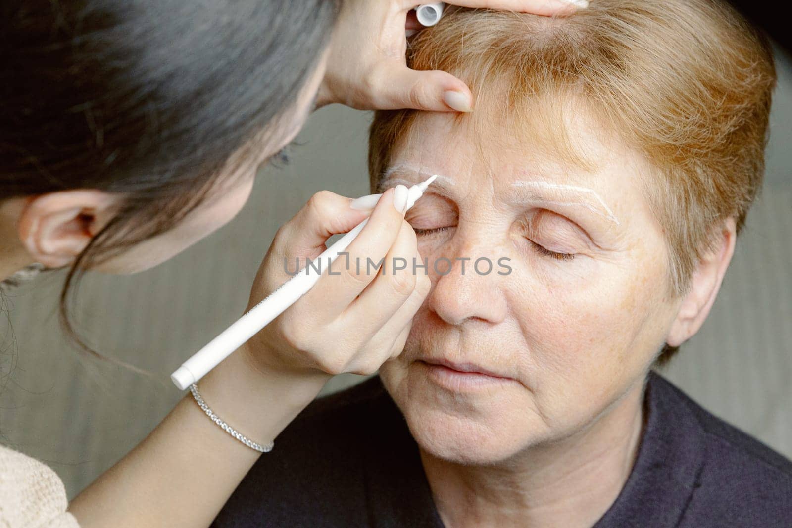 One young Caucasian girl cosmetologist draws a contour with a white marker on the right eyebrow of an elderly beautiful woman with her eyes closed, sitting in a home beauty salon, top side close-up view.