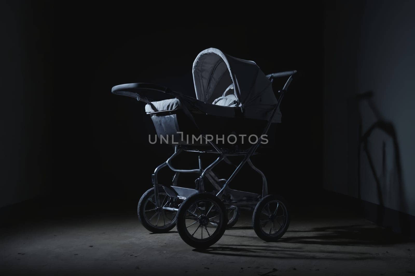 Bathed in a spotlight's cold gleam against an otherwise empty wall, stands an antique baby stroller. This black and white image captures the eerie loneliness and nostalgic overtones imprinted in its silhouette and the surrounding quiet.