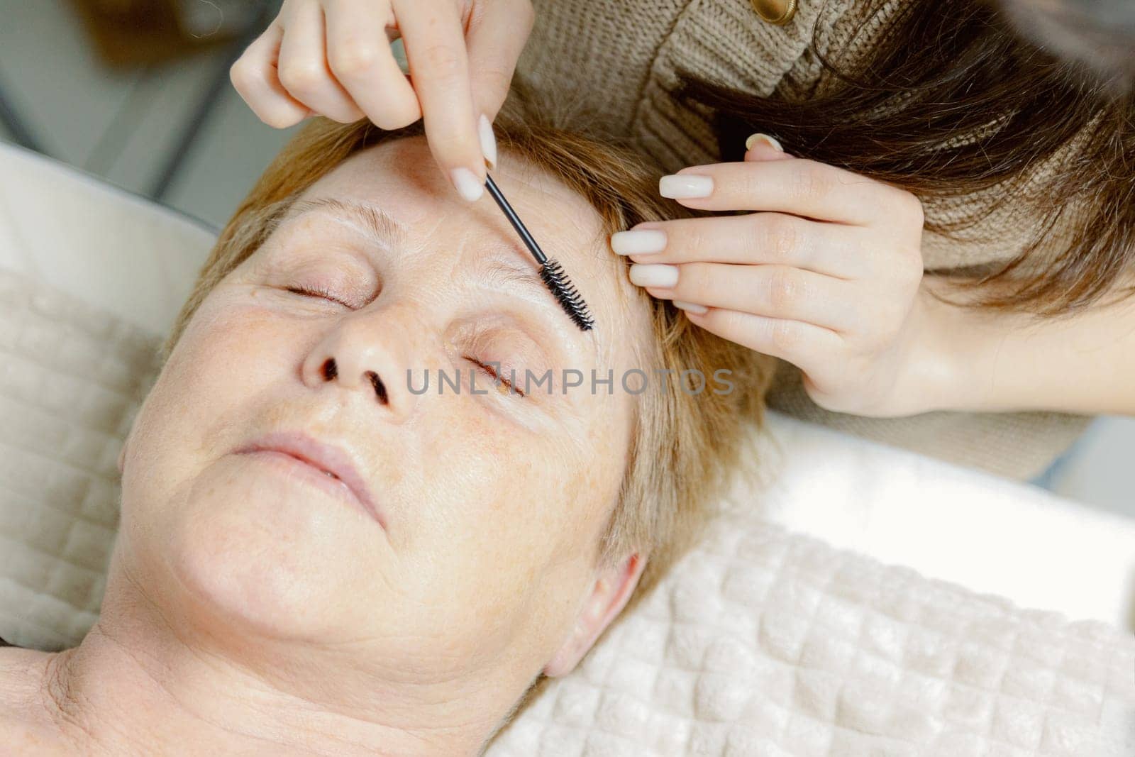 One young Caucasian brunette young girl cosmetologist combs the eyebrows of an elderly woman with closed eyes using a round brush lying on a cosmetology bed in a home beauty salon, top side close-up view.