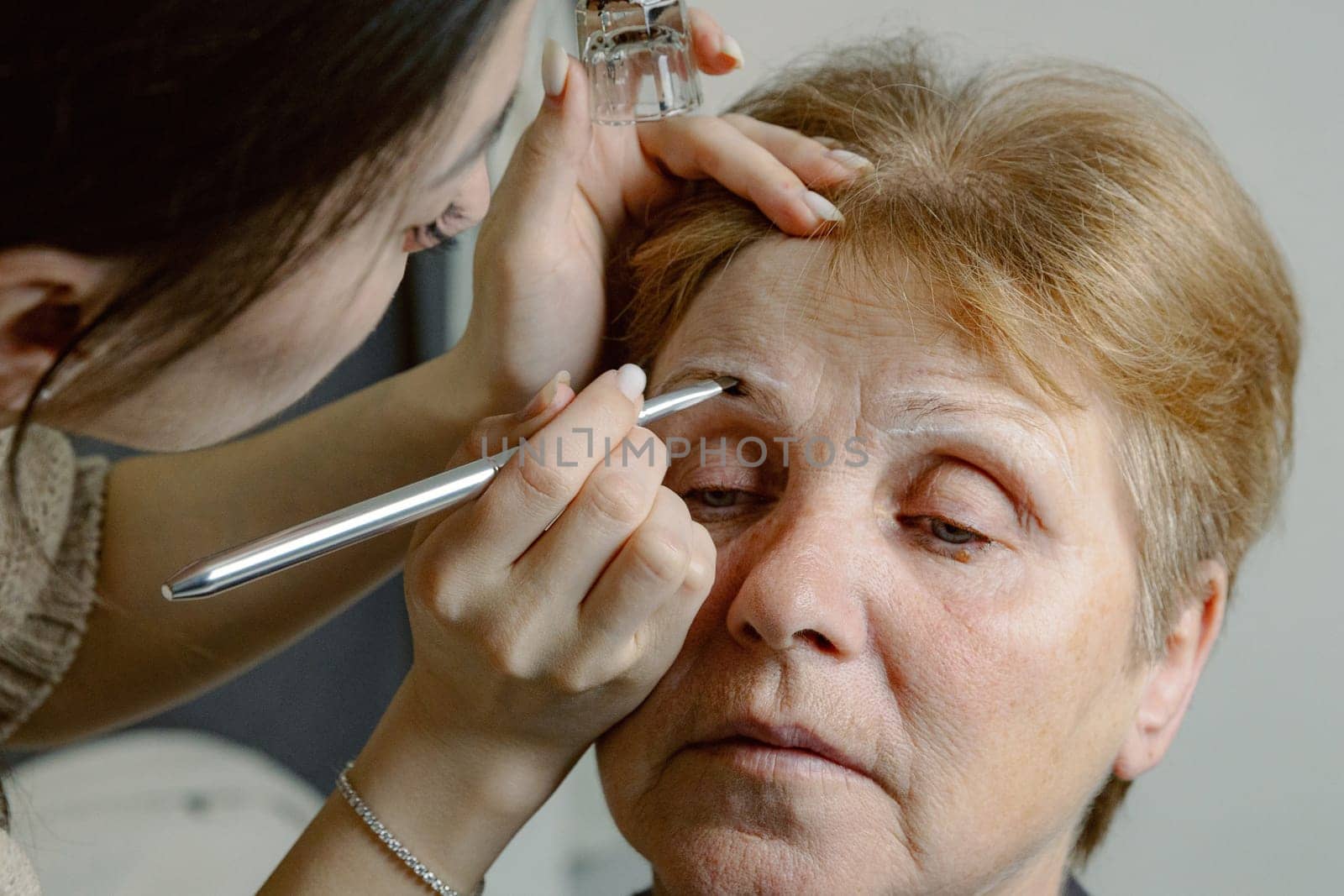 A cosmetologist paints the eyebrows of an elderly woman. by Nataliya