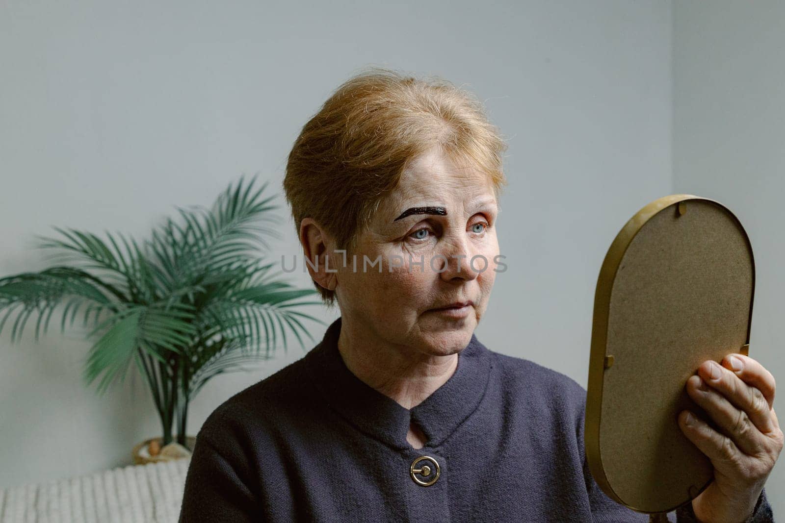 One young Caucasian elderly woman looks in the mirror, carefully examining her right eyebrow painted with dark brown paint, sitting in a home beauty salon on a cosmetology bed, close-up side view.