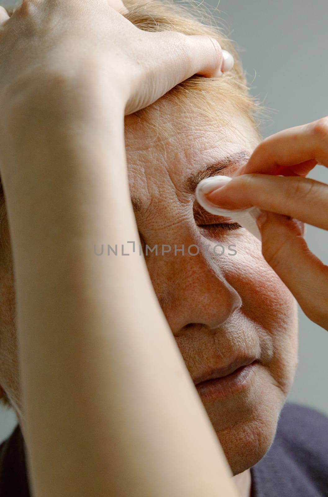 One young Caucasian young unrecognizable girl cosmetologist carefully removes the remaining paint from the left eyebrow of an elderly woman with closed eyes with a cotton pad, using a cleanser, holding her head with her hand, in a home beauty salon, side view close-up.