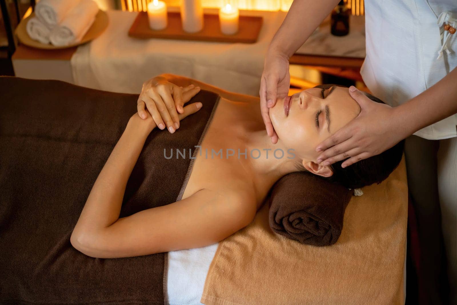 Caucasian woman enjoying relaxing anti-stress head massage and pampering facial beauty skin recreation leisure in warm candle lighting ambient salon spa in luxury resort or hotel. Quiescent